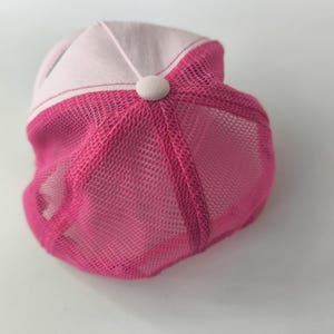 May include: A baseball cap featuring a light pink front panel and a vibrant hot pink mesh back. The cap includes a matching fabric-covered button at the center top where the panels meet, showcasing a structured design with breathable mesh sections.