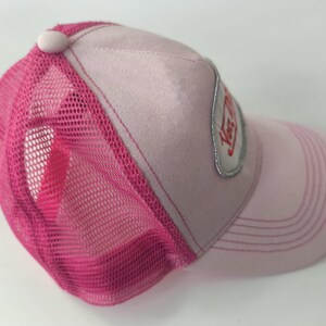 May include: A light pink trucker hat featuring a bright fuchsia mesh back and a matching pink button on the crown. The front panel displays a silver-bordered patch with red text. The curved brim has stitched detailing in a matching pink tone.