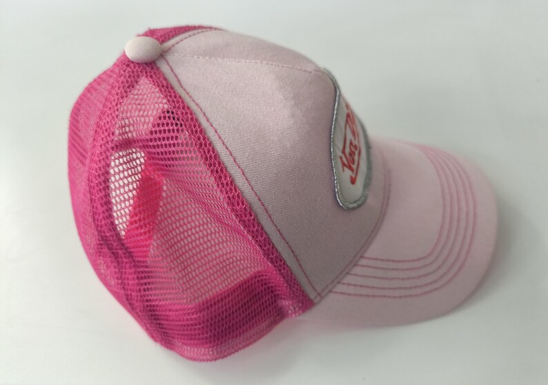 May include: A light pink trucker hat featuring a bright fuchsia mesh back and a matching pink button on the crown. The front panel displays a silver-bordered patch with red text. The curved brim has stitched detailing in a matching pink tone.