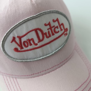 May include: A light pink baseball cap featuring an oval-shaped patch on the front. The patch has a white background with a silver-grey embroidered border and the text Von Dutch in red embroidery. The cap has visible stitching along the brim.