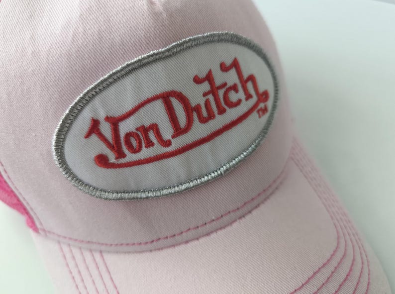 May include: A light pink baseball cap featuring an oval-shaped patch on the front. The patch has a white background with a silver-grey embroidered border and the text Von Dutch in red embroidery. The cap has visible stitching along the brim.