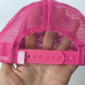 May include: A bright pink trucker hat featuring a mesh back panel and a plastic snapback closure. The cap includes a small rectangular fabric tag attached to the side of the adjustable strap, which is also in a matching shade of vibrant pink.
