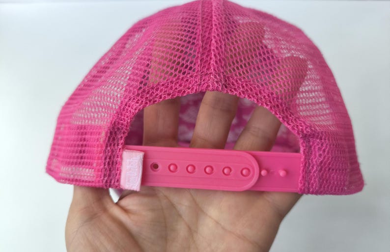 May include: A bright pink trucker hat featuring a mesh back panel and a plastic snapback closure. The cap includes a small rectangular fabric tag attached to the side of the adjustable strap, which is also in a matching shade of vibrant pink.