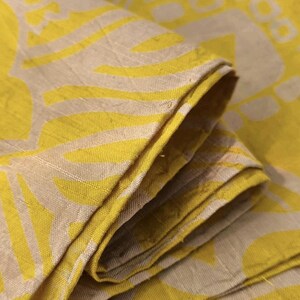 May include: A folded piece of fabric featuring a bright yellow and beige pattern. The textile displays a repeating geometric design with bold yellow shapes set against a neutral beige background, showing a visible woven texture throughout the material.
