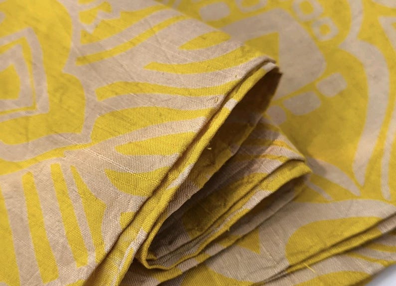May include: A folded piece of fabric featuring a bright yellow and beige pattern. The textile displays a repeating geometric design with bold yellow shapes set against a neutral beige background, showing a visible woven texture throughout the material.