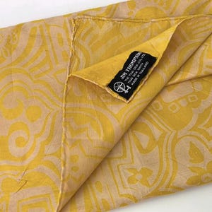 May include: A yellow and beige patterned silk scarf featuring a black fabric label that reads JIM THOMPSON THE THAI SILK CO. LTD. THAI SILK 100% MADE IN THAILAND with a small circular logo depicting a spinning wheel and thread.