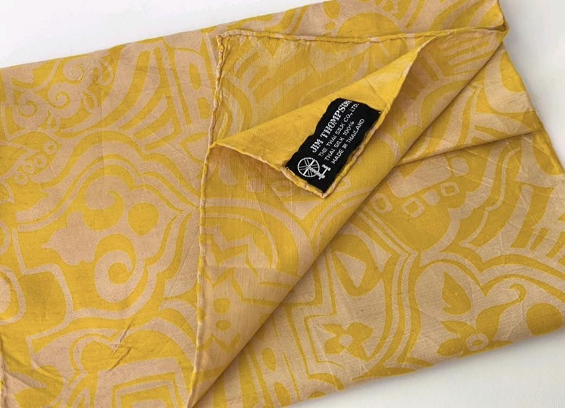 May include: A yellow and beige patterned silk scarf featuring a black fabric label that reads JIM THOMPSON THE THAI SILK CO. LTD. THAI SILK 100% MADE IN THAILAND with a small circular logo depicting a spinning wheel and thread.