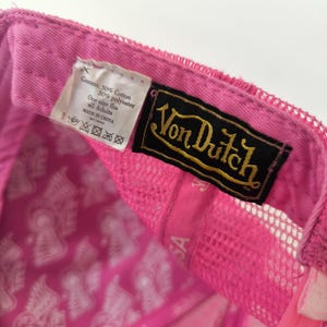 May include: A bright pink trucker hat featuring a black and gold Von Dutch brand label sewn inside. The interior lining is printed with a repeating white graphic pattern. A white fabric tag displays content information and care instructions.