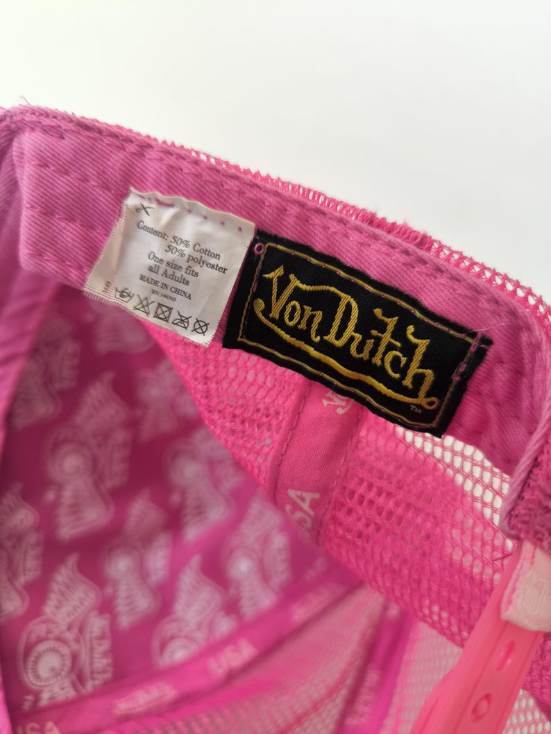 May include: A bright pink trucker hat featuring a black and gold Von Dutch brand label sewn inside. The interior lining is printed with a repeating white graphic pattern. A white fabric tag displays content information and care instructions.