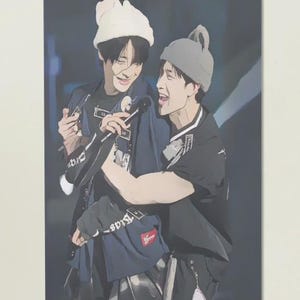 Könnte beinhalten: Illustration von zwei Personen mit Mützen und dunkler Kleidung, die sich umarmen und lächeln. Sie halten Mikrofone, was auf eine Aufführung oder Veranstaltung hindeutet. Der Hintergrund ist ein verschwommenes, dunkles Blau.