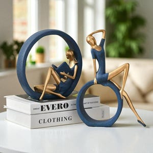 Könnte beinhalten: Zwei dekorative Skulpturen mit goldenen und blauen Figuren. Eine Figur sitzt in einem blauen Ring und liest ein Buch. Die andere Figur sitzt auf einem blauen Ring, einen Arm erhoben. Die Skulpturen stehen auf einer weißen Oberfläche.