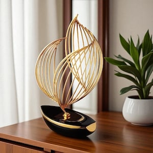 Puede incluir: Escultura decorativa de metal dorado con forma de velero estilizado. La base es negra con detalles dorados, y la vela es de alambre dorado. La escultura está sobre una superficie de madera. En el fondo, una planta en maceta blanca.