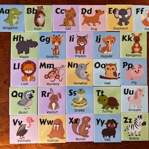 May include: Alphabet flashcards featuring colorful illustrations of animals, each paired with a corresponding letter. The cards include an alligator, bear, cat, dog, elephant, fox, hippo, giraffe, impala, jaguar, kangaroo, lion, monkey, narwhal, owl, pig, quail, rabbit, snake, turtle, unicorn, vulture, walrus, xerus, yak, and zebra.