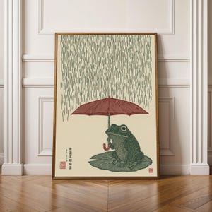 Peut inclure: Impression encadrée d'une grenouille verte sous un parapluie rouge, assise sur une feuille de nénuphar. Des lignes verticales symbolisent la pluie. L'œuvre comprend du texte japonais et un sceau rouge.