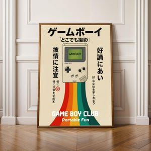 Op de afbeelding: Een ingelijste poster met een retro-ontwerp met een Game Boy-console en Japanse tekst. De Game Boy heeft een regenboogpad en de tekst "GAME BOY CLUB Portable Fun" staat onderaan. De poster heeft een vintage esthetiek.