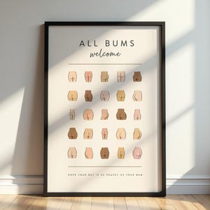 Op de afbeelding: Een ingelijste wandprint met een crèmekleurige achtergrond met de tekst ALL BUMS welcome bovenaan en HOPE YOUR DAY IS AS PEACHY AS YOUR BUM onderaan. Het midden toont een raster van vijfentwintig geïllustreerde menselijke billen in diverse huidtinten.