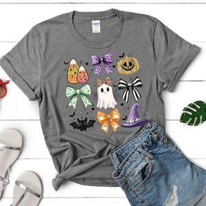 May include: A gray t-shirt featuring a Halloween-themed design. The design includes candy corn, bows, a jack-o'-lantern, a ghost, a witch's hat, and a bat. The shirt is paired with denim shorts.
