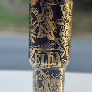 May include: Black and gold insulated tumbler featuring artwork from "The Legend of Zelda: Tears of the Kingdom." The design includes a warrior on horseback, the game's title, and other detailed illustrations. The tumbler has a gold rim and lid.