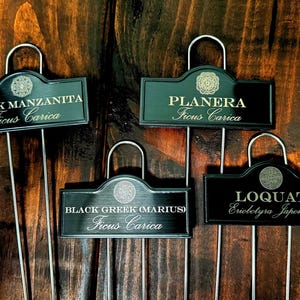 May include: Four black rectangular plant markers with silver metal stakes. Each marker features a unique circular emblem at the top and white text identifying plant varieties: Black Manzanita, Planera, Black Greek (Marius), and Loquat.