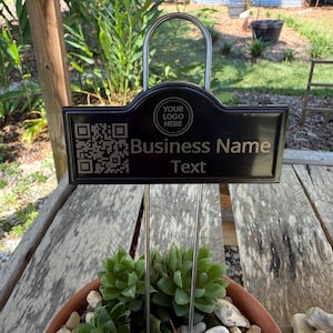 May include: Black metal sign with a curved top, featuring a QR code and the words "Business Name" and "Text". The sign is held by a metal arch and is placed above a potted succulent plant.