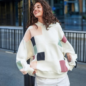 Könnte beinhalten: Ein cremeweißer Kapuzenpullover mit Patchwork-Design, bestehend aus quadratischen Einsätzen in gedeckten Tönen wie Pfirsich, Salbeigrün, Anthrazit und Altrosa. Der langärmelige Pullover verfügt über eine Kapuze und gerippte Bündchen.