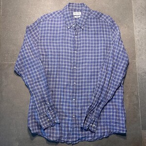 May include: A blue and white plaid button-down shirt with long sleeves. The shirt has a button-down collar and buttoned cuffs. The shirt is made of a lightweight fabric and is laid flat on a gray surface.