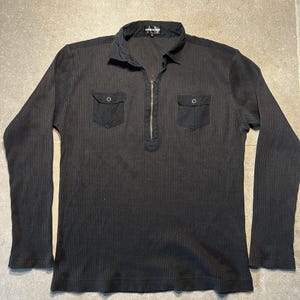 May include: Black long-sleeve shirt with a ribbed texture. The shirt features a collared neckline, a quarter-zip closure, and two small chest pockets with button closures. The shirt has a simple, classic design.