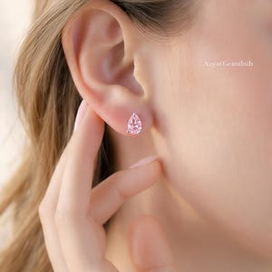 May include: A pair of pink, teardrop-shaped stud earrings. The earrings are set in a silver metal and are worn on the ear. The earrings are a light pink colour and are being touched by a hand. The text "Aayat Gemshub" is in the background.