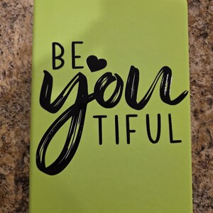 May include: A lime green notebook with the words "BE YOU TIFUL" in black script. The word "you" is stylized with a heart replacing the "o". The notebook has a smooth, matte finish.