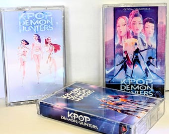 May include: Three clear plastic cassette cases featuring K-Pop Demon Hunters official soundtrack artwork. Two cases stand upright, one displays three figures in white outfits, the other shows five figures in dark gear. One case lies flat with group art.