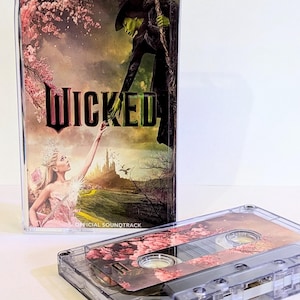 May include: A clear plastic cassette case featuring the Wicked Official Soundtrack cover art with a green-skinned figure and a blonde figure reaching toward each other, alongside a matching cassette tape with a floral cherry blossom design.