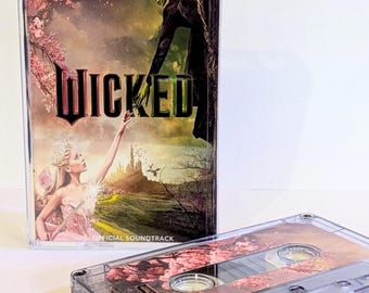 May include: A clear plastic cassette case featuring the Wicked Official Soundtrack cover art with a green-skinned figure and a blonde figure reaching toward each other, alongside a matching cassette tape with a floral cherry blossom design.