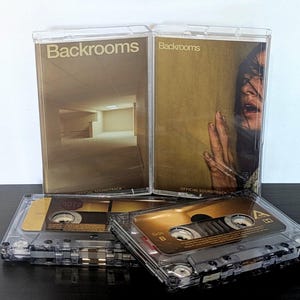 Peut inclure: Deux boîtiers de cassette en plastique transparent avec le titre Backrooms et le texte Official Soundtrack. Un boîtier montre un couloir vide aux tons jaunes, l'autre un visage et une main. Deux cassettes aux étiquettes dorées sont devant.