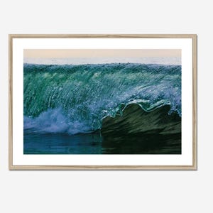 May include: A framed print featuring a close-up of a breaking ocean wave. The wave is a mix of deep teal and blue hues, with white foam and spray. The artwork is encased in a light-colored wooden frame, creating a serene coastal theme.