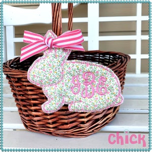 May include: A pink and white floral fabric bunny with a pink monogram and a pink and white striped bow. The bunny is attached to a brown wicker basket.