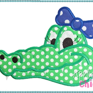 May include: Green alligator head with white polka dots and a blue bow with white polka dots. The alligator has big eyes and a friendly smile. The text "Applique Chick" is in the lower right corner of the image.