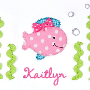May include: A pink fish with white polka dots and a pink bow, surrounded by green seaweed and gray bubbles. The name "Kaitlyn" is embroidered below the fish.