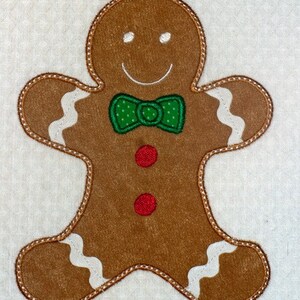 May include: A brown gingerbread man with a smiling face, white eyes, and a green polka-dot bow tie. The gingerbread man has two red buttons and white icing details. The edges are outlined with a brown stitch.
