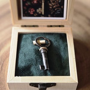 May include: Open wooden box with a hinged lid, containing a vintage key on a green velvet base. The box has a small framed picture of a key on the inside of the lid. The key is dark metal with a decorative handle.