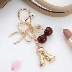Può includere: Un portachiavi color oro con un fiocco, due ciliegie e il ciondolo con la lettera "A". Le ciliegie sono di colore rosso scuro. Il portachiavi è attaccato a un fermaglio dorato.