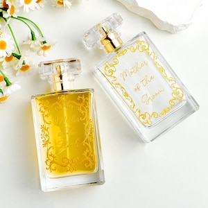 Op de afbeelding: Twee rechthoekige glazen parfumflesjes met gouden verstuivers en doorzichtige doppen. De ene fles bevat amberkleurige vloeistof en heeft de gouden tekst Mother of the Bride. De andere is leeg met de gouden tekst Mother of the Groom.