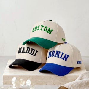 Può includere: Tre cappellini da baseball con corona color bianco sporco e visiere colorate. Il cappellino in alto ha una visiera verde e la scritta CUSTOM in verde. Quello a sinistra ha una visiera nera e la scritta MADDI in nero. Quello a destra ha una visiera blu e la scritta NONIN in blu.