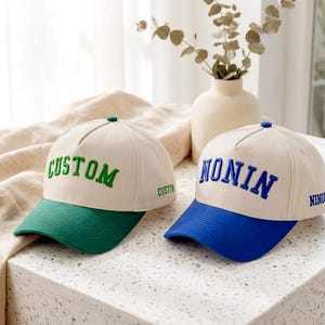 Può includere: Due cappellini da baseball con corona color crema e visiera colorata. Il cappellino a sinistra ha una visiera verde e la scritta ricamata in verde CUSTOM sul davanti e sul lato. Il cappellino a destra ha una visiera blu e la scritta NONIN in blu.