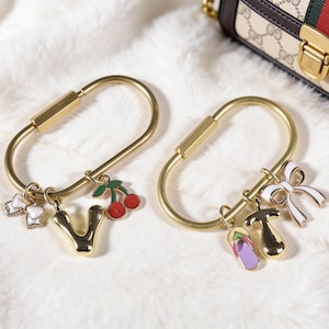 Può includere: Due portachiavi color oro con ciondoli decorativi. Un portachiavi presenta un ciondolo a forma di lettera "V", un ciondolo a ciliegia e un ciondolo a cuore. L'altro ha un ciondolo a fiocco, un ciondolo a infradito e un ciondolo a forma di T.