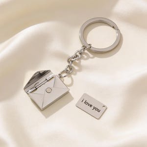 Könnte beinhalten: Silberfarbener Schlüsselanhänger mit einem kleinen, aufklappbaren Umschlag. Im Inneren befindet sich ein kleines Schild mit der Aufschrift "i love you". Der Schlüsselanhänger ist an einer Kette und einem großen Ring befestigt.