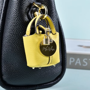 May include: A yellow miniature handbag charm with a gold-colored circular tag and a small padlock. The tag has the inscription "My Way" in cursive. The charm is attached to a black leather handbag with a gold-colored chain.