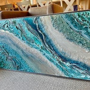 May include: Abstract ocean-themed artwork in a rectangular silver frame. The piece features textured layers of teal, turquoise, and white resin, creating a dynamic, wave-like effect. White lines meander across the surface, enhancing the water's flow.