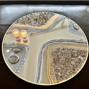 May include: A round decorative mirror with a gold-colored frame. The mirror's surface features a unique design with embedded crystals and a layered pattern in shades of gold, gray, and white. Two lit candles are reflected in the mirror.