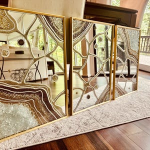 May include: A set of three rectangular wall mirrors framed in gold, featuring decorative geode-inspired accents. The surfaces are adorned with textured layers of crushed glass, glitter, and metallic sand in shades of gold, bronze, and white.