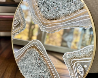 May include: Round decorative mirror with a gold-colored metal frame. The mirror's surface is embellished with a geode-inspired design, featuring layers of silver, gold, and white accents, and embedded with sparkling crystals.